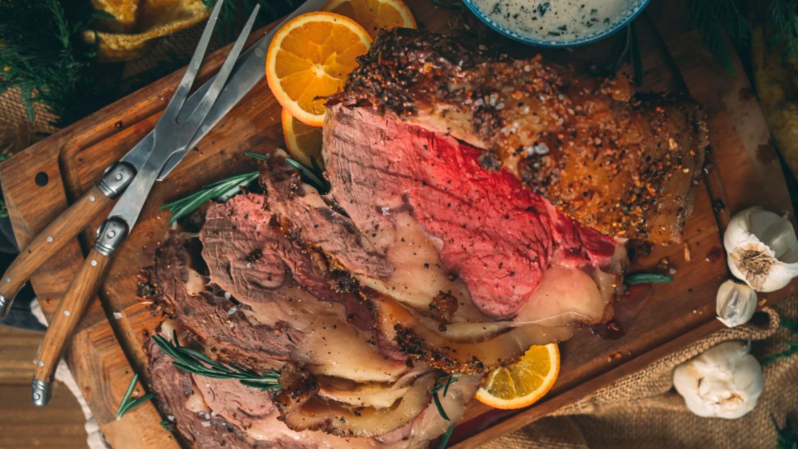 Sliced prime rib roast on a wooden board with rosemary, orange slices, and a fork and knife set.