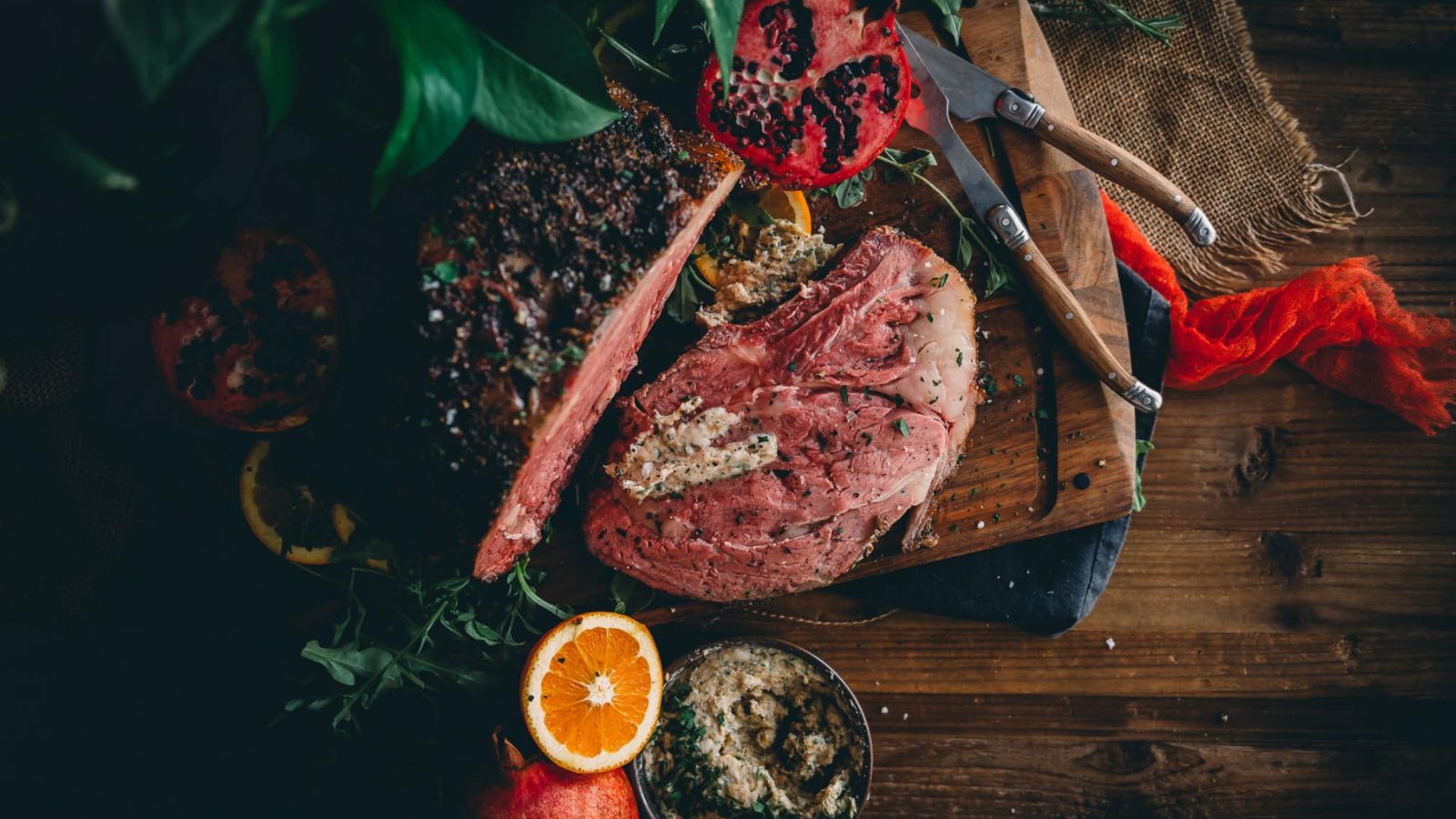 Sliced roast beef on a wooden board with herbs, a pomegranate, and an orange. Two knives are placed beside it.