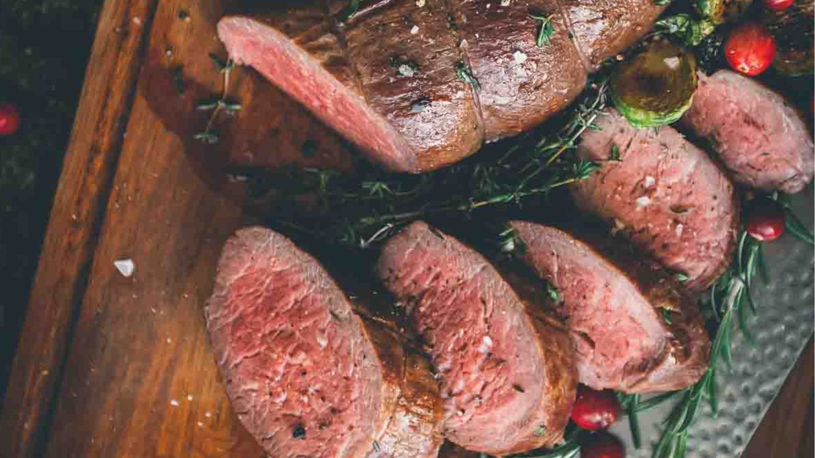 Sliced medium-rare beef roast on a wooden cutting board, garnished with sprigs of herbs and whole peppercorns, tempts the palate as you ponder the best sauces for beef tenderloin.