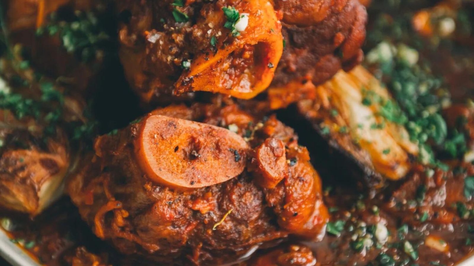 Close-up of a dish featuring cooked meat with a bone, garnished with chopped herbs in a rich sauce.