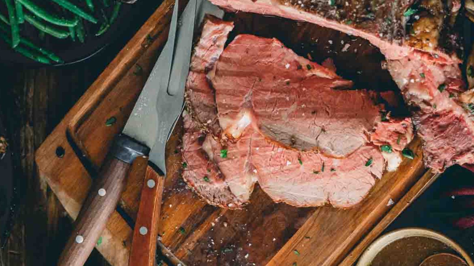 Sliced roast beef on a wooden cutting board with a knife and a bowl of green beans nearby.