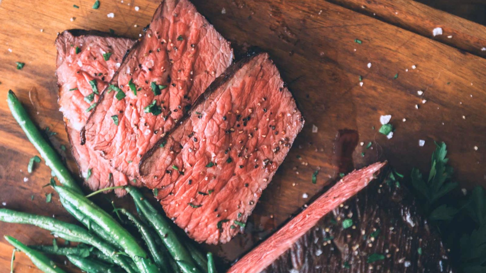 Slices of medium-rare steak garnished with herbs next to green beans on a wooden board.