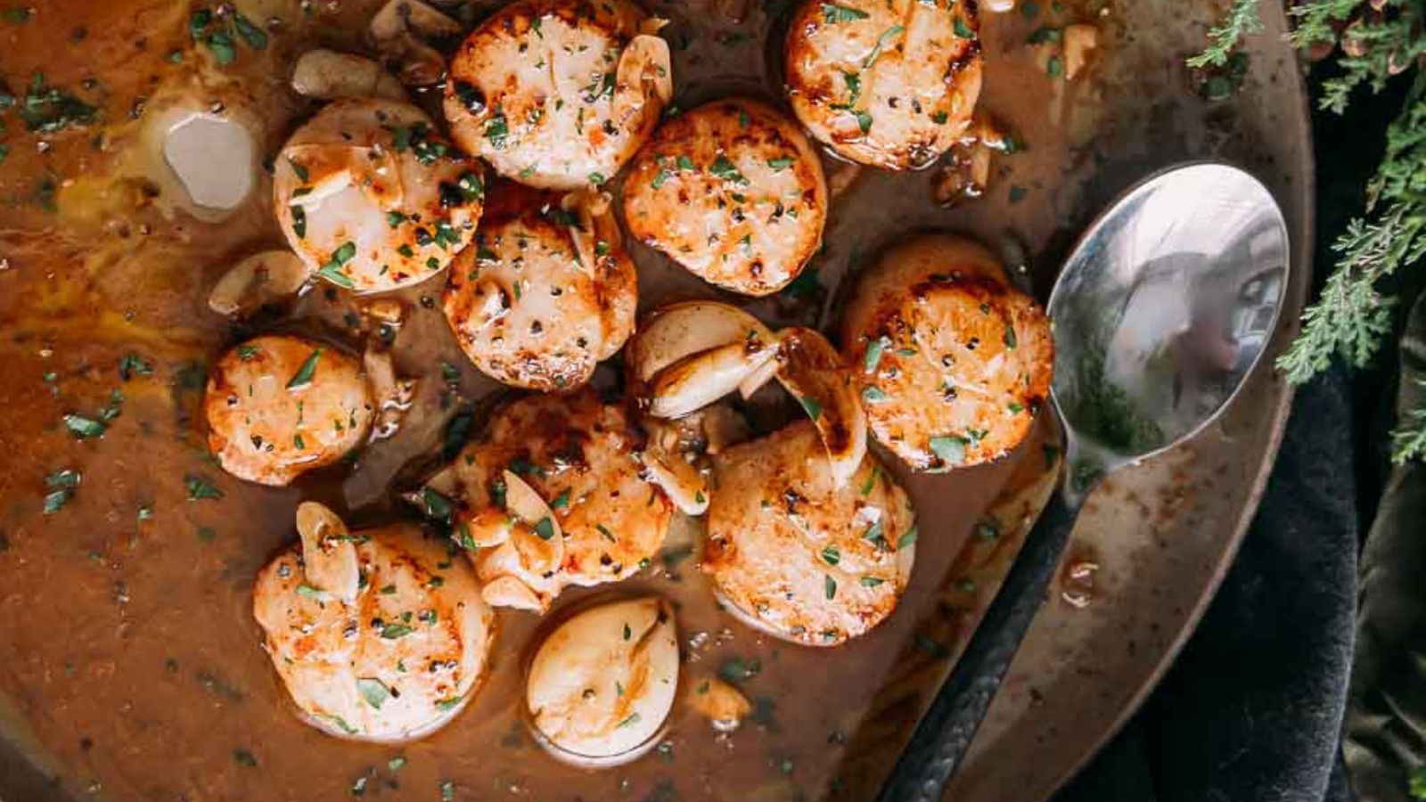 Pan-seared scallops with garlic and herbs on a plate, accompanied by a spoon.