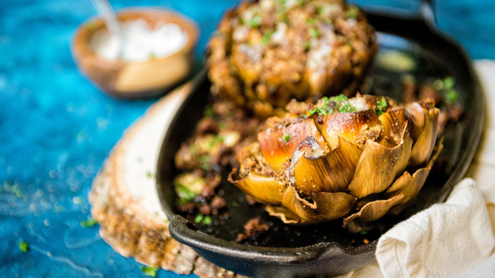 Two roasted artichokes on a black skillet, garnished with herbs. A small bowl with a spoon is in the background on a textured blue surface.