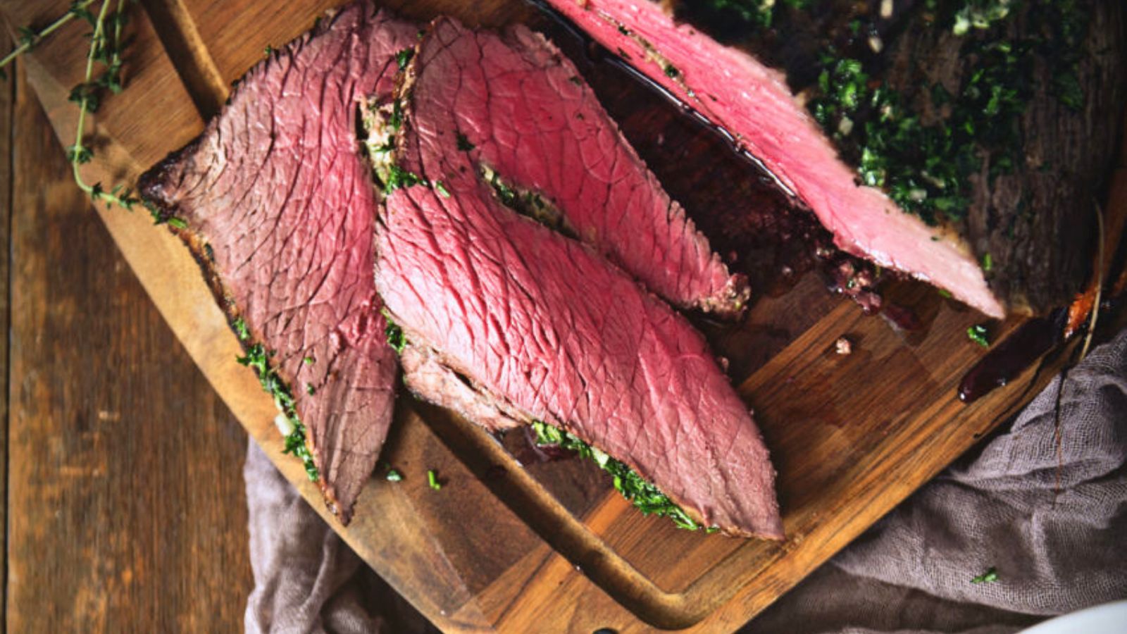 Sliced roast beef on a wooden cutting board, garnished with chopped herbs.