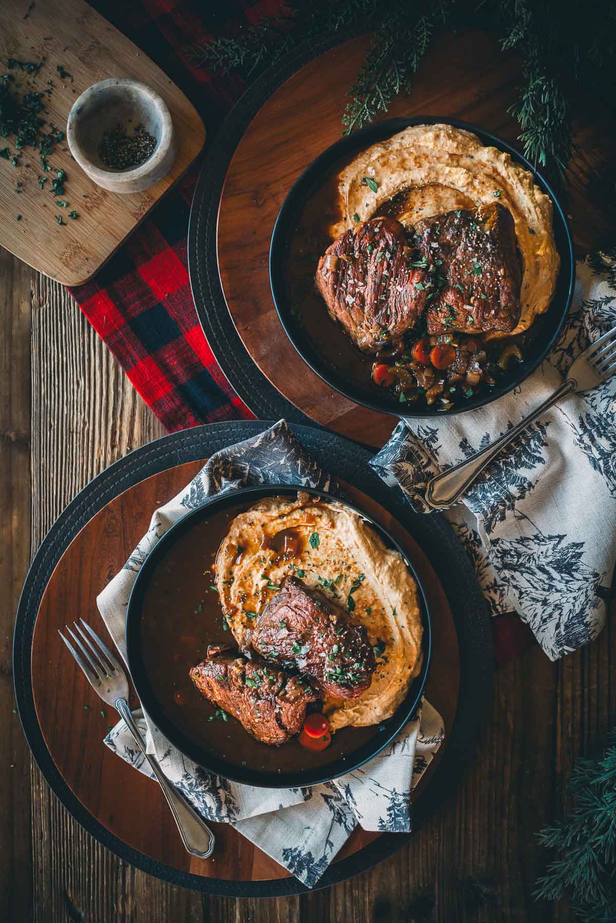 Two plates of sous vide short ribs with mashed root veggies and vegetables on a wooden table with decorative napkins and a plaid cloth.