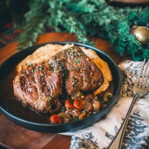 A plate with two pieces of sous vide short ribs on a bed of vegetables, garnished with herbs, next to a fork and a decorative napkin.