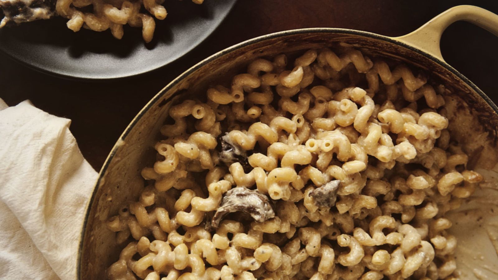 A pot of macaroni and cheese with mushrooms. A cloth napkin is beside it, and a black plate with a small serving can be seen at the edge.