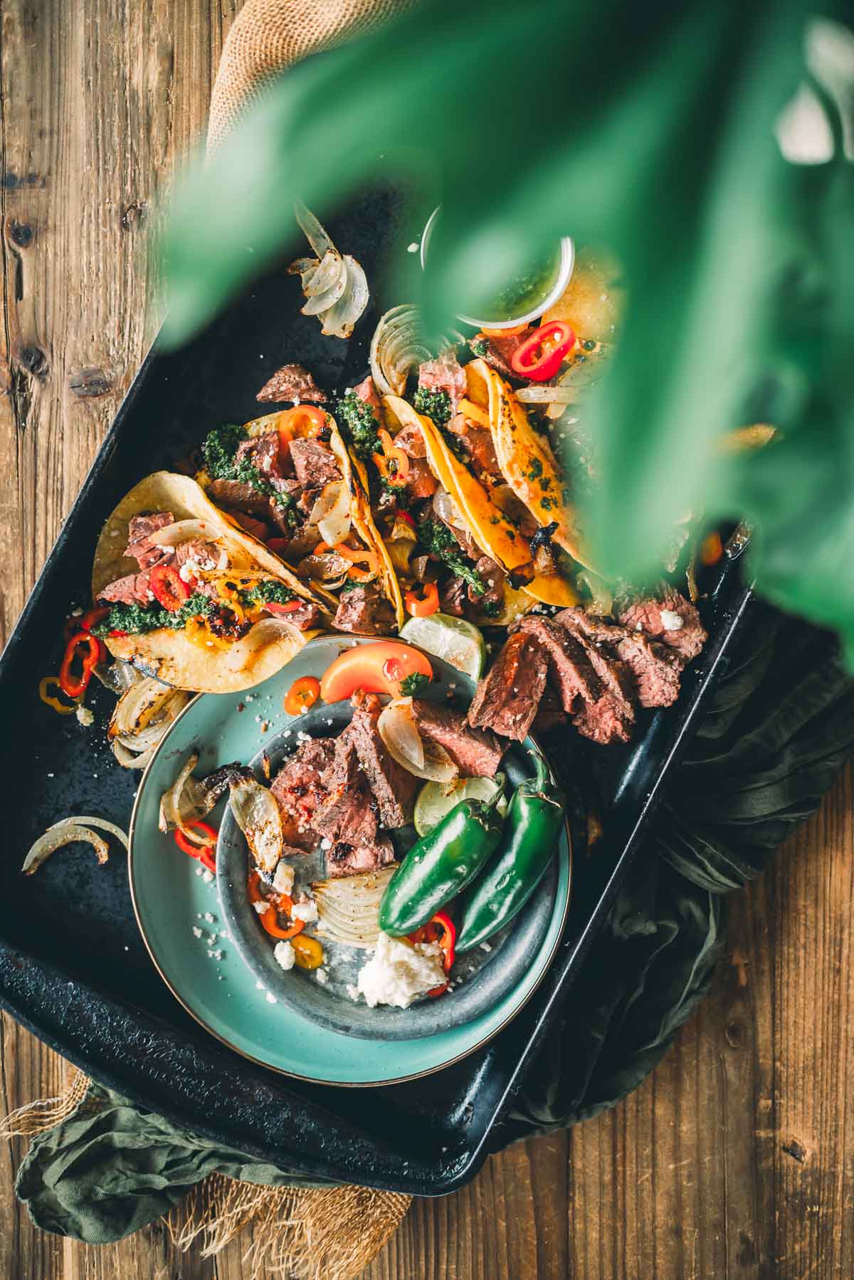 Tacos with sliced hanger steak, chilies, onions, and herbs on a dark platter, garnished with lemon slices. A green plant leaf in the foreground.