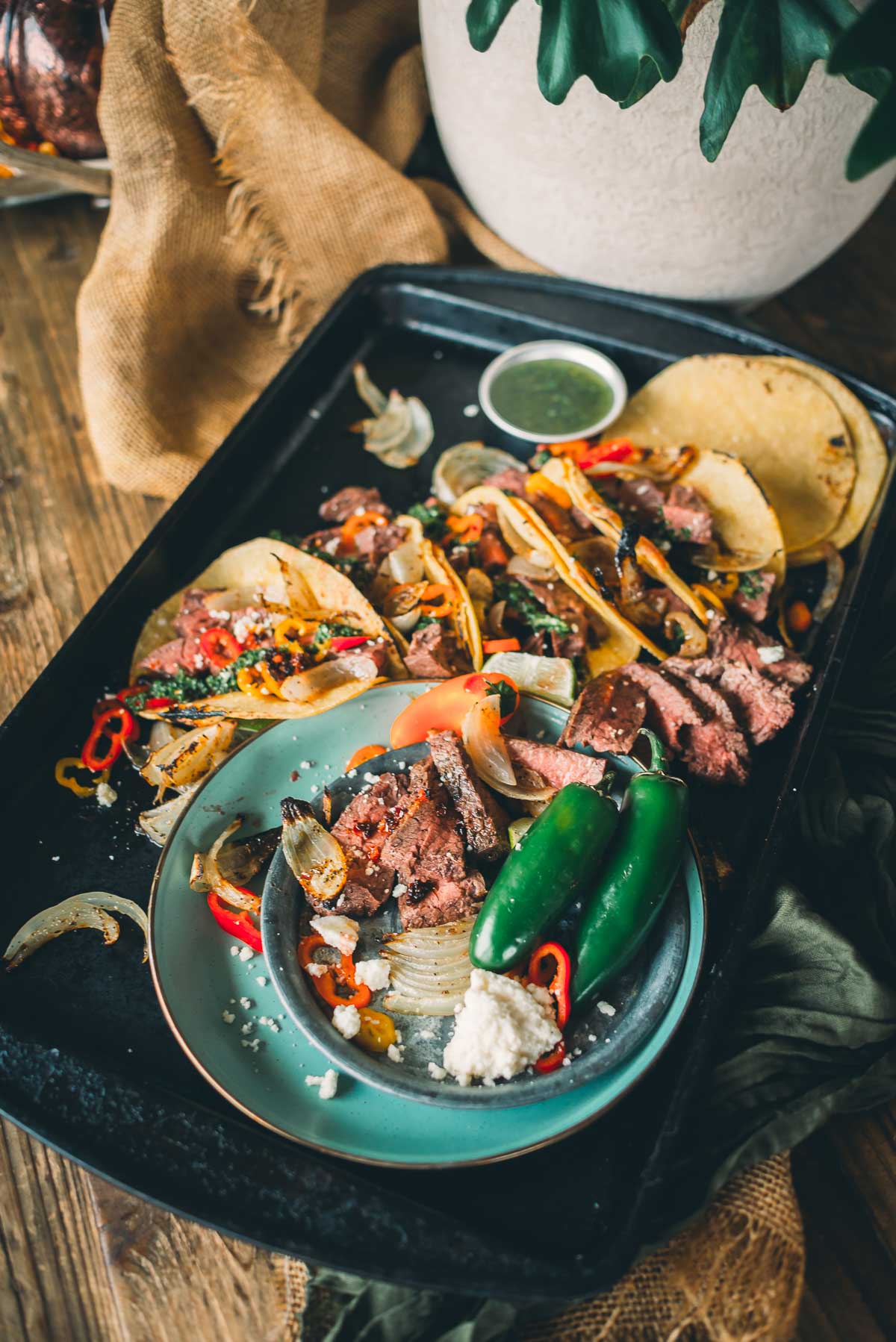 A plate with sliced hanger steak tacos, jalapeños, and cheese, surrounded by taco toppings on a tray. Garnished with onions and peppers, with a small bowl of chimichurri.