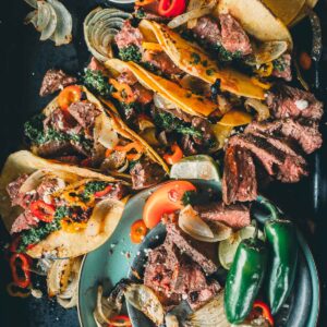 Tacos filled with broiled hanger steak steak, onions, and peppers on a plate, with jalapeños, cheese, and a cup of chimichurri on the side.