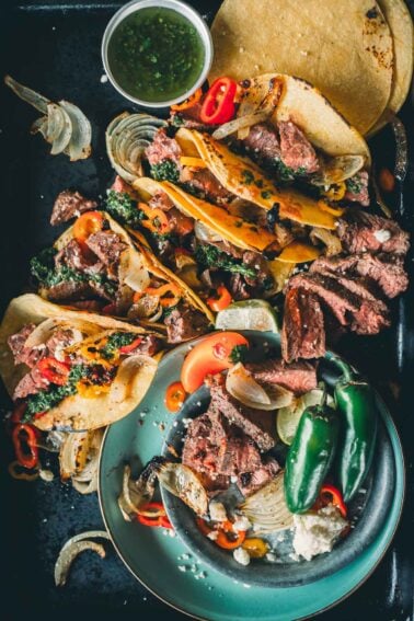 Tacos filled with broiled hanger steak steak, onions, and peppers on a plate, with jalapeños, cheese, and a cup of chimichurri on the side.