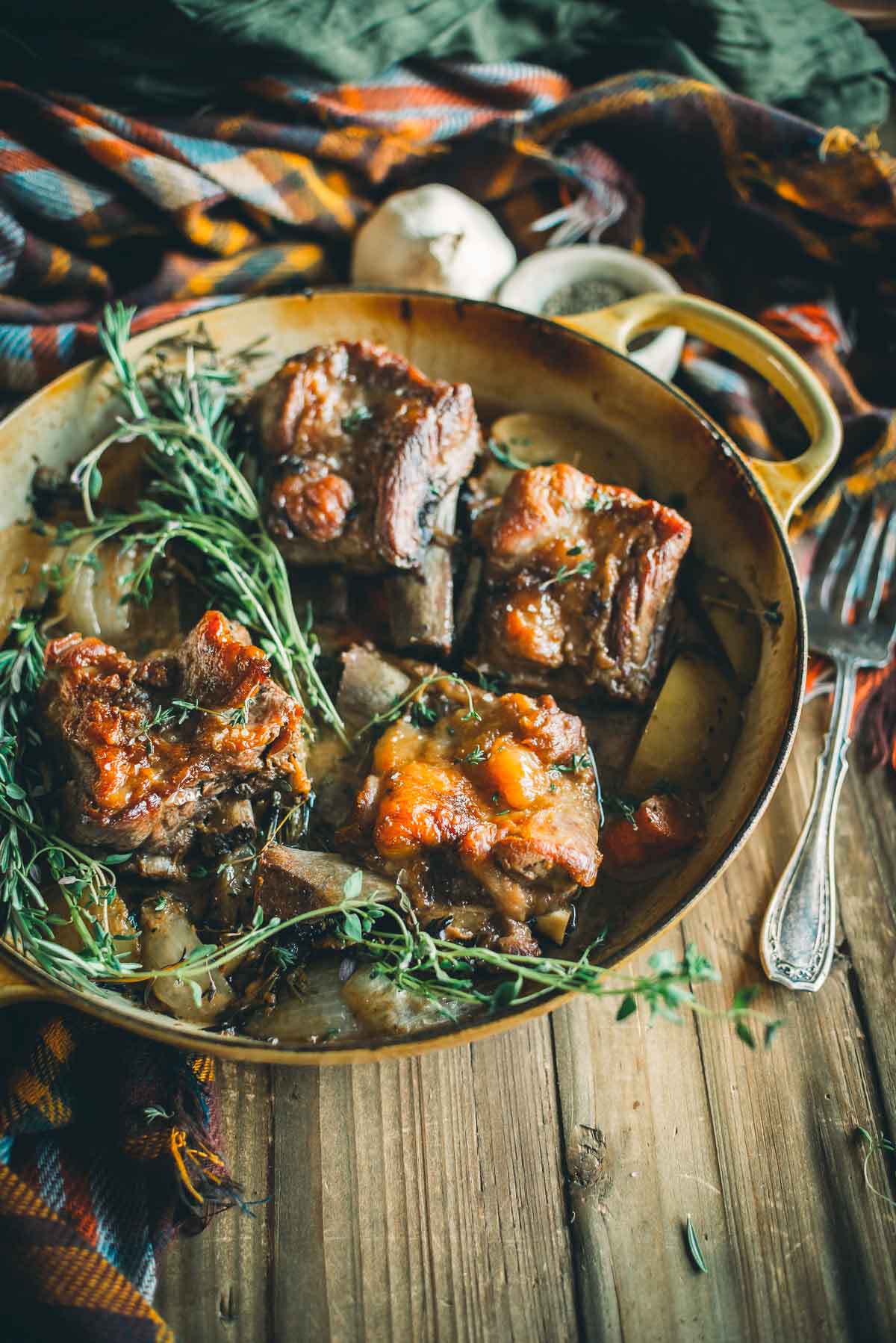 Braised pork short ribs in a pan, garnished with fresh herbs, set on a wooden table with a plaid cloth and a fork.