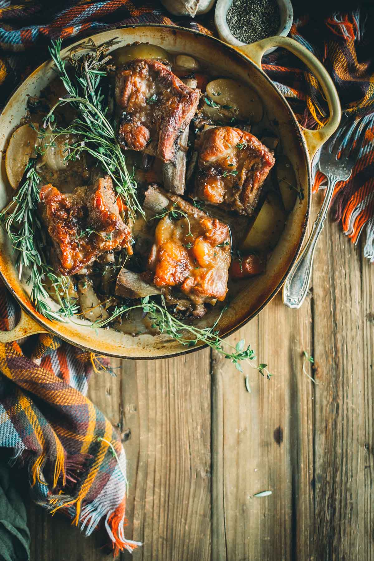 A rustic dish of pork short ribs pieces on a wooden table, garnished with fresh herbs. A colorful cloth and utensils are beside the dish.