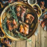 A rustic dish with braised pork short ribs on a platter, garnished with rosemary. Garlic and a small bowl of pepper are on the side. A colorful cloth is draped in the background.