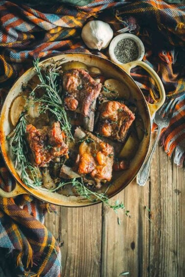 A rustic dish with braised pork short ribs on a platter, garnished with rosemary. Garlic and a small bowl of pepper are on the side. A colorful cloth is draped in the background.