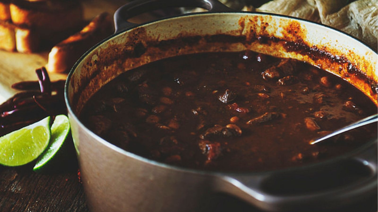 A pot of rich, dark chili containing beans and meat, with a metallic ladle. Lime wedges and dried chilies are visible in the background.