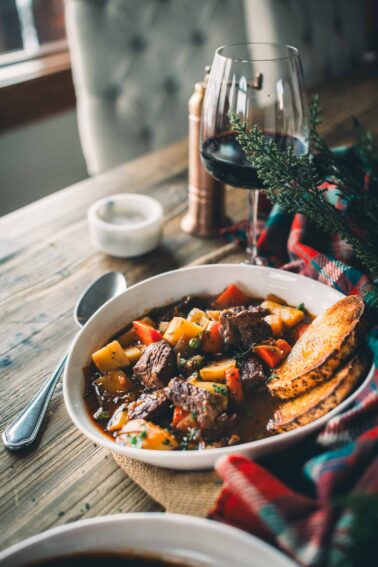 A bowl of Guinness beef stew with vegetables and bread slices, set on a wooden table with a glass of red wine, salt shaker, and a plaid cloth nearby.