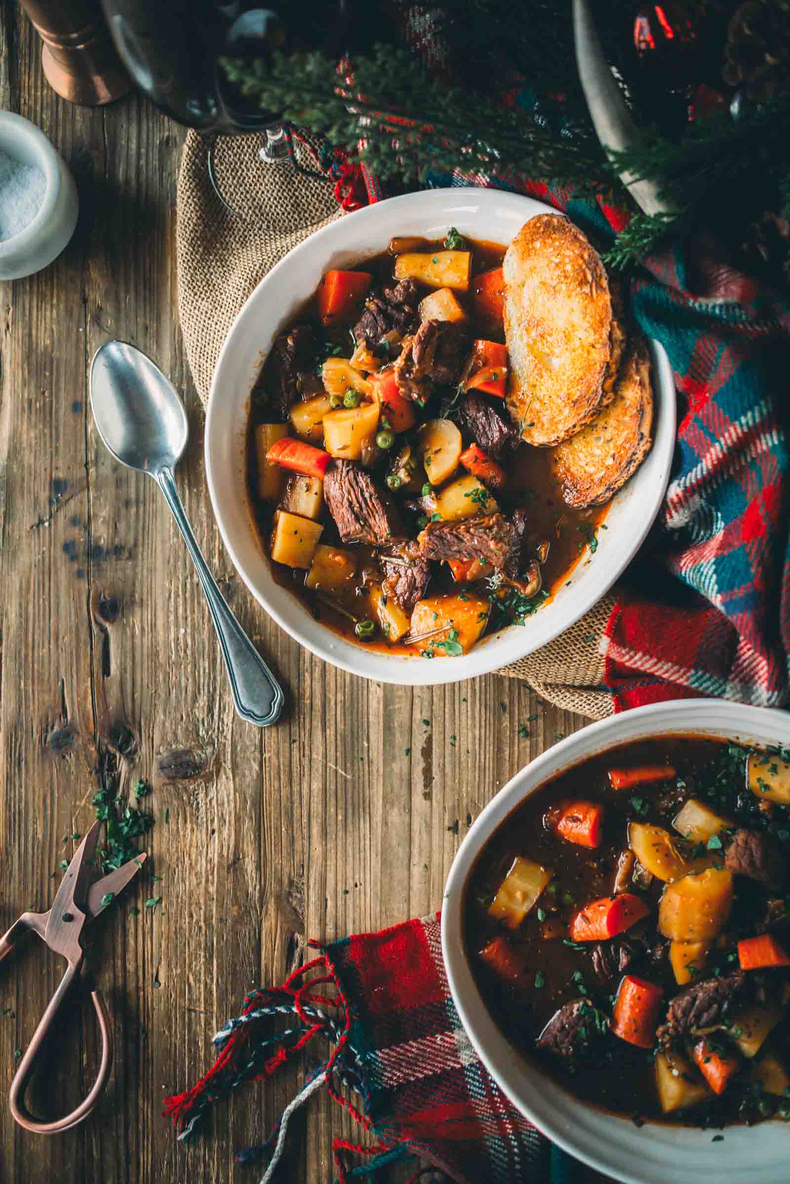 A bowl of Guinness beef stew with vegetables and bread on a wooden table, garnished with herbs. A spoon and scissors are nearby, along with a plaid cloth for decoration.