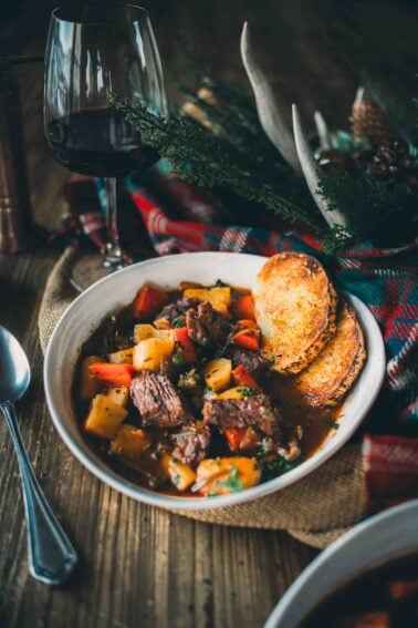 A bowl of Guinness beef stew with vegetables and toasted bread slices, next to a glass of red wine on a wooden table with plaid fabric accents.