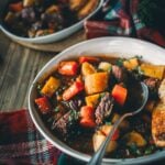 Two bowls of Guinness beef stew with carrots and potatoes, garnished with herbs, placed on a wooden table with a red plaid cloth. A spoon rests in the front bowl beside toasted bread slices.