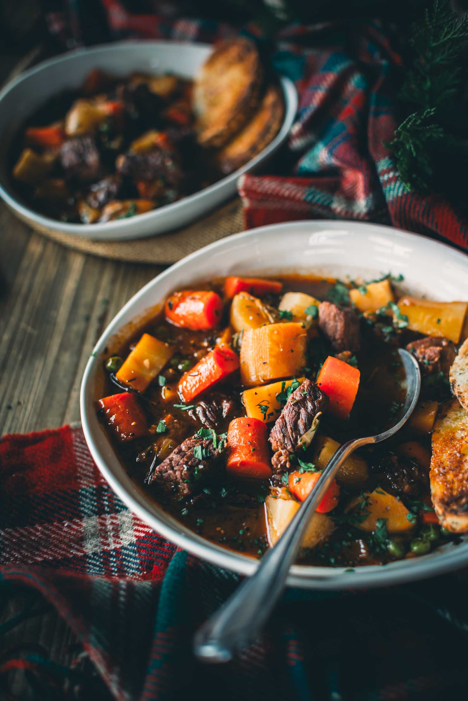 Two bowls of Guinness beef stew with carrots and potatoes, garnished with herbs, placed on a wooden table with a red plaid cloth. A spoon rests in the front bowl beside toasted bread slices.