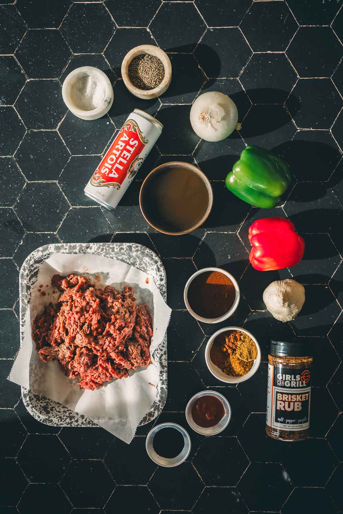 Ingredients for cooking venison chili: ground deer meat, bowls of spices, brisket rub, garlic, onions, bell peppers, a can of tomatoes, and a bowl of broth on a black hexagonal tile surface.