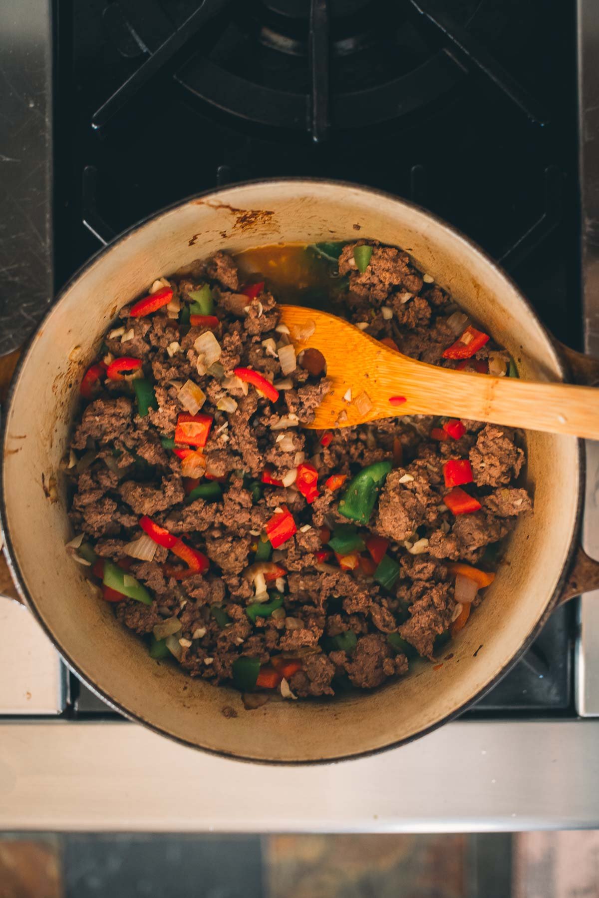 A pot on the stove containing browned minced venison, chopped green and red bell peppers, and onions, stirred with a wooden spoon.