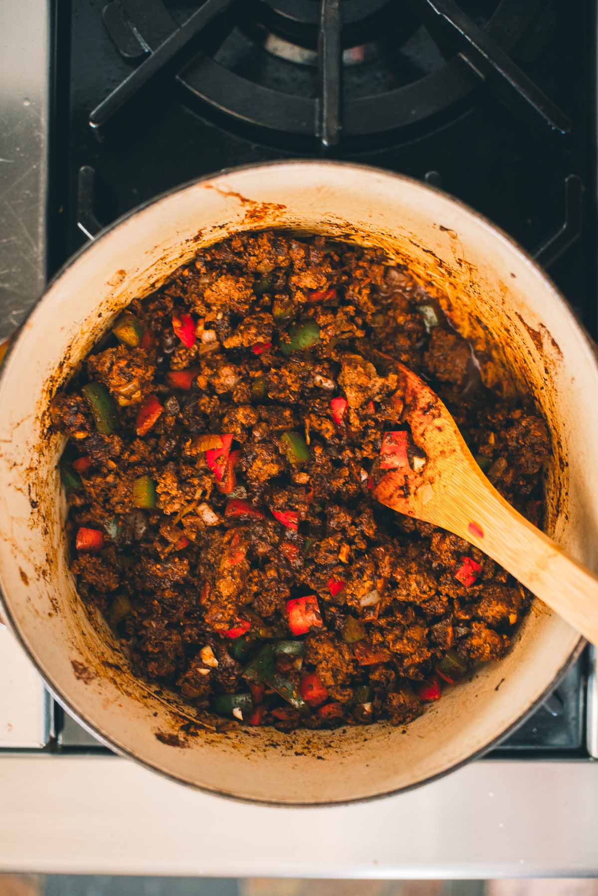 A pot of chili with ground deer meat, red and green peppers, and seasonings, being stirred with a wooden spoon on a stovetop.