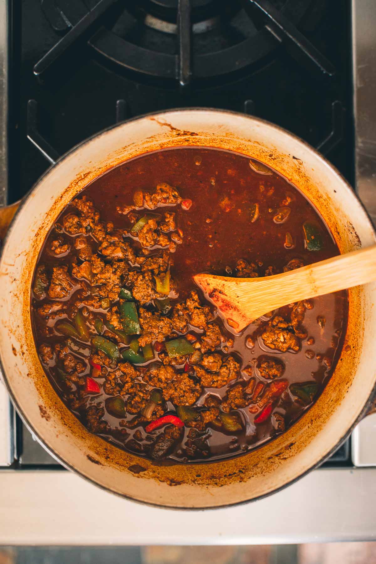 A pot of ground venison chili with ground meat, green and red peppers, and a wooden spoon on a stovetop.