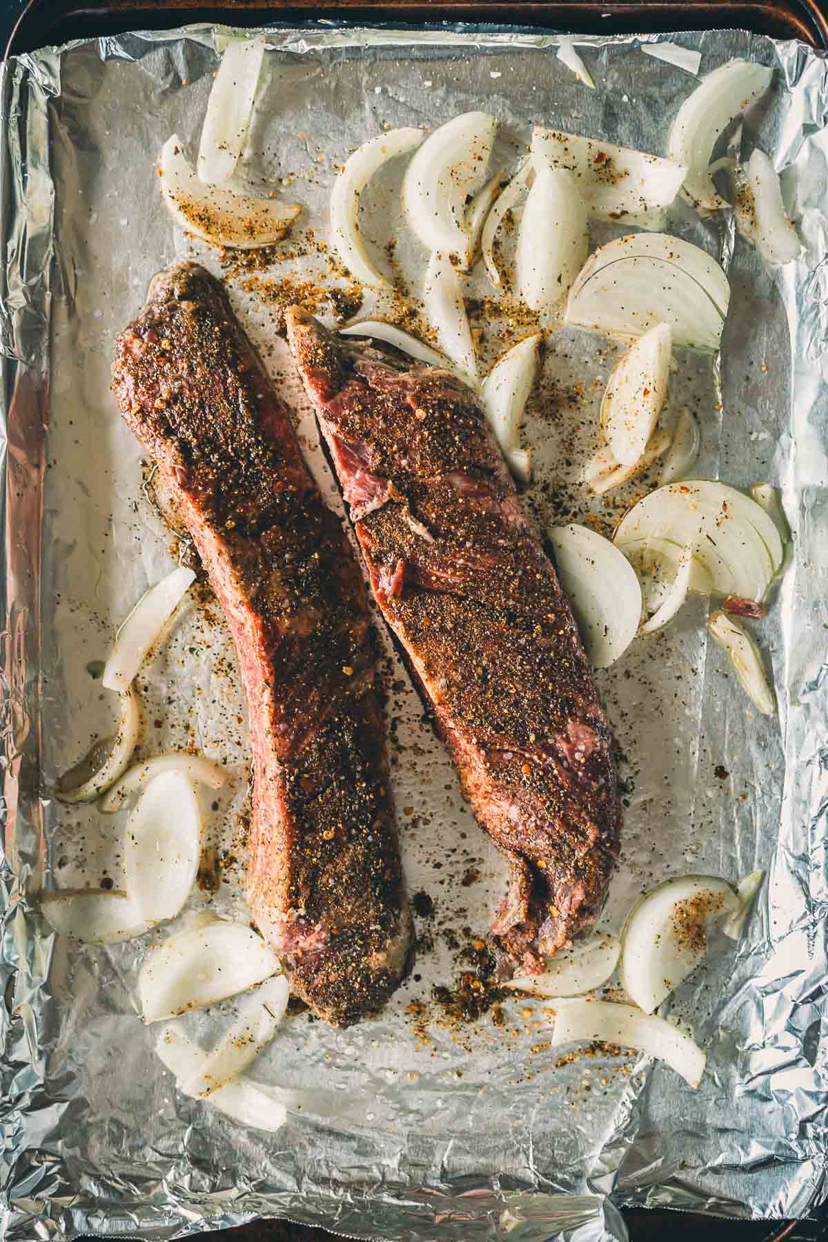 Two spiced beef hanger steaks and sliced onions on a foil-lined baking sheet.