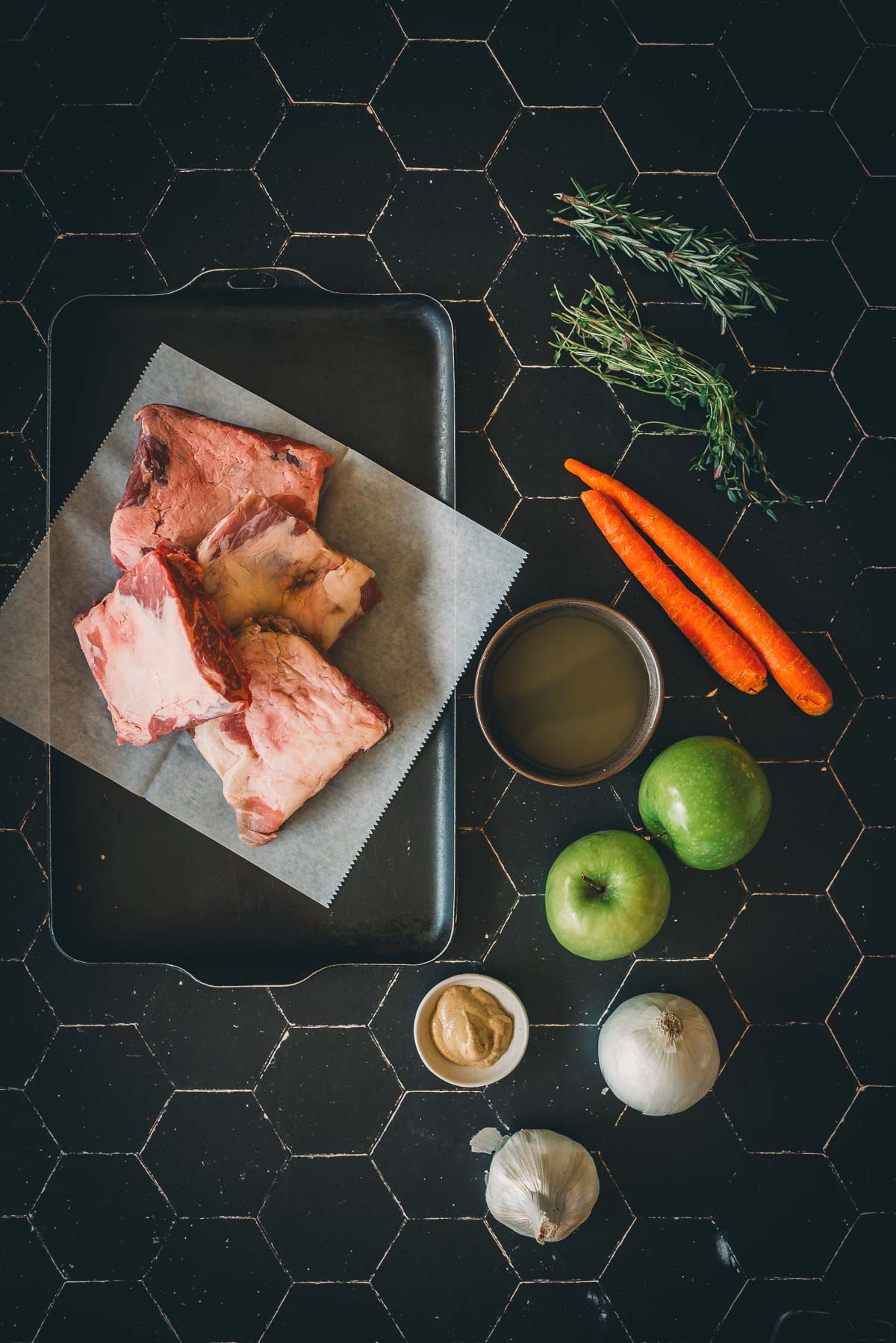 Top-down view of the ingredients for cider braised pork short ribs; raw slabs of pork on a tray with parchment paper, surrounded by carrots, rosemary, a bowl of broth, two green apples, an onion, garlic, and a small bowl of mustard on a hexagonal tile surface.