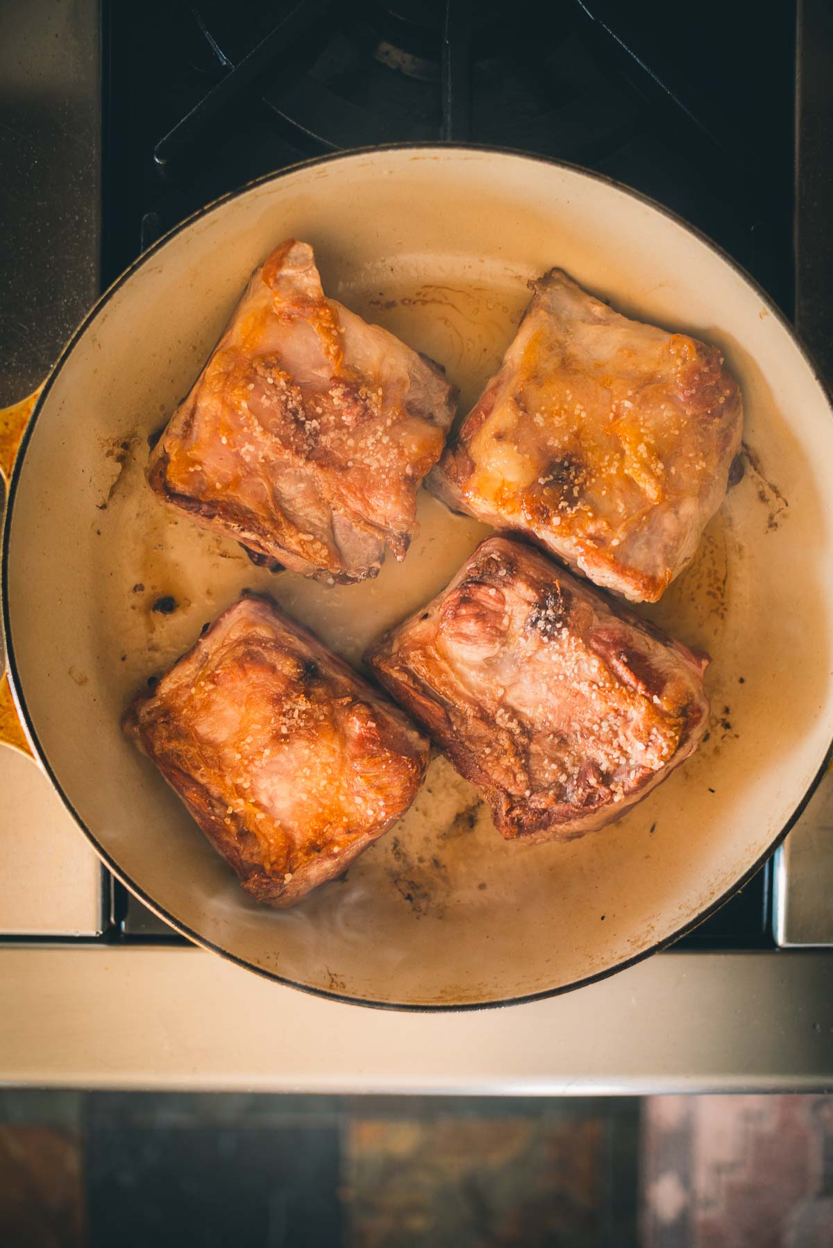 Four salted slabs of pork short ribs are cooking in a round pan over a stovetop.