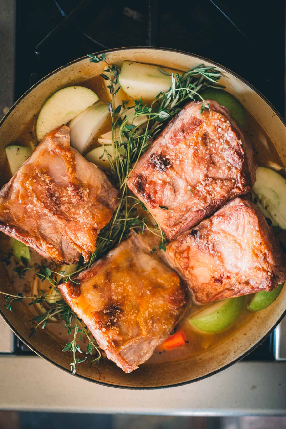 Browned pork short ribs in a pan with sliced apples, onions, and sprigs of thyme.