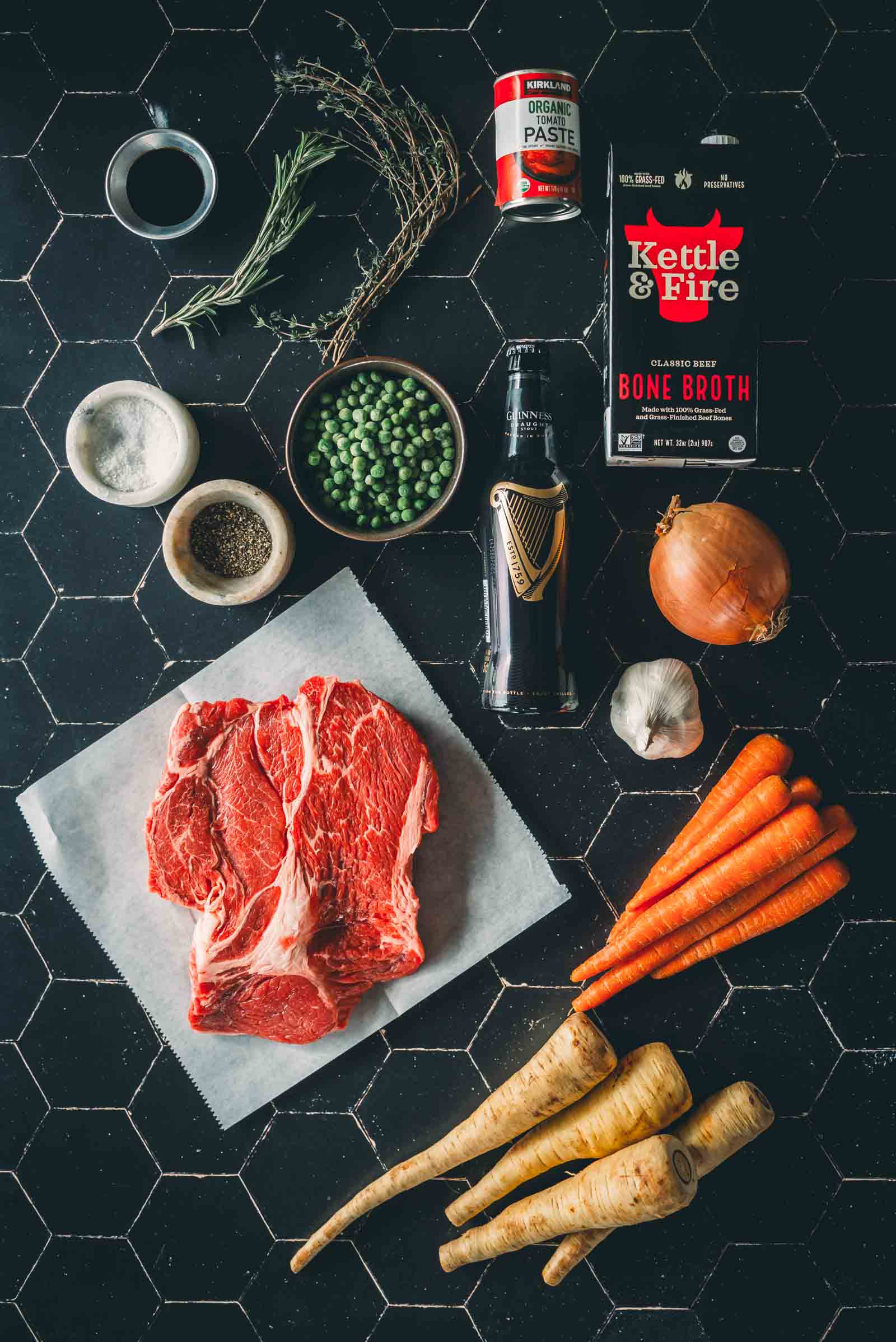 Raw beef chuck roast, carrots, parsnips, onion, garlic, a can of tomato paste, bone broth, peas, rosemary, salt, and pepper are arranged on a dark tile surface.