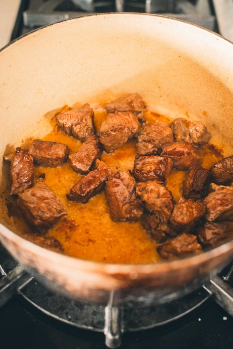 Cubed beef browning in a pot on a stovetop, with visible caramelization and some liquid at the bottom.