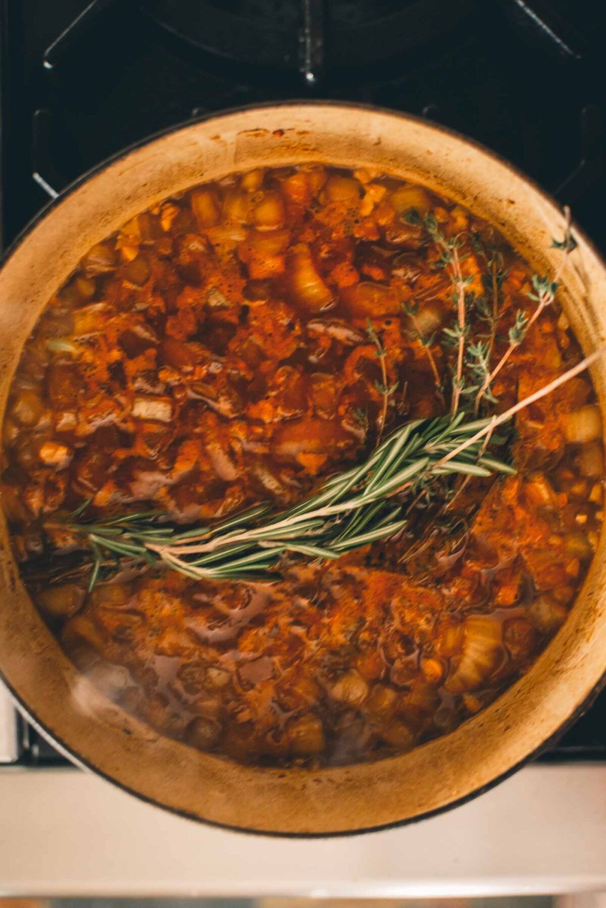 A pot of stew simmering on a stove, containing diced vegetables, herbs, and a reddish broth with whole sprigs of rosemary and thyme on top.