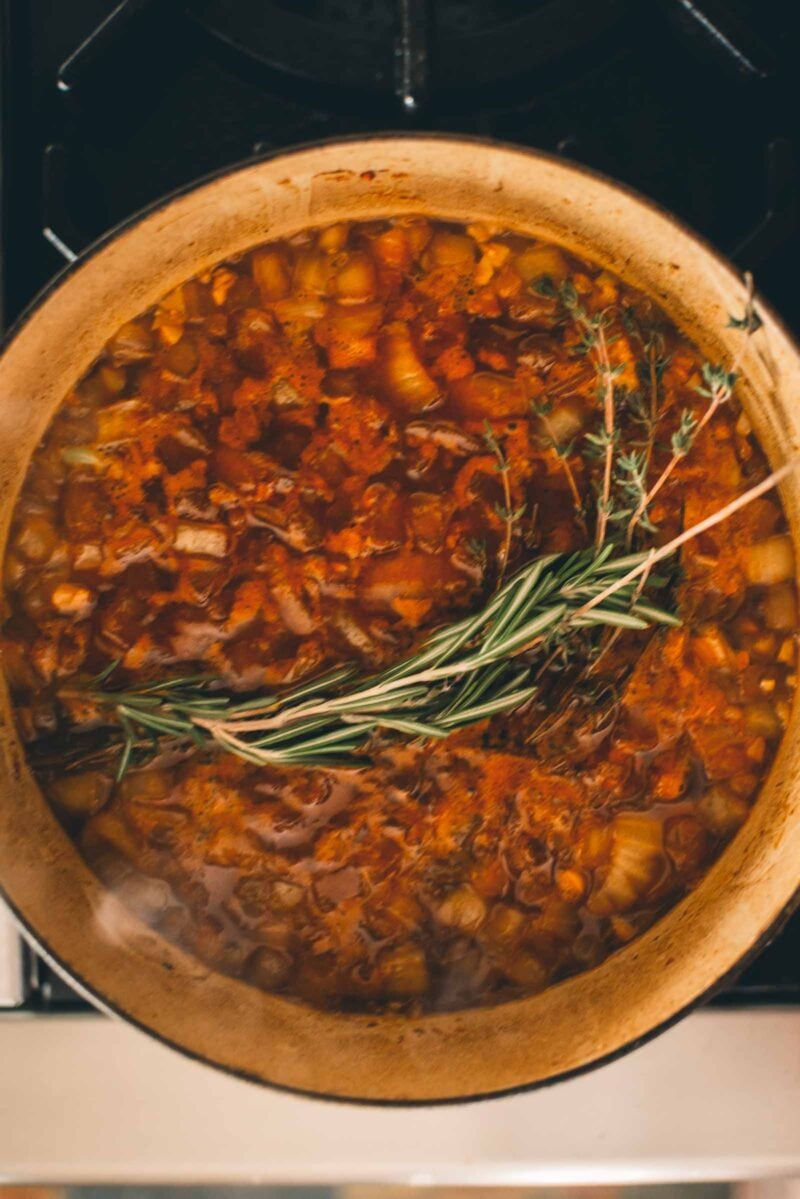 A pot of stew simmering on a stove, containing diced vegetables, herbs, and a reddish broth with whole sprigs of rosemary and thyme on top.