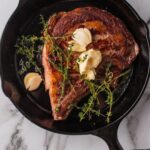 A cast iron skillet contains a seared piece of meat topped with butter and garnished with thyme. Text overlay promotes a guide on searing meat.