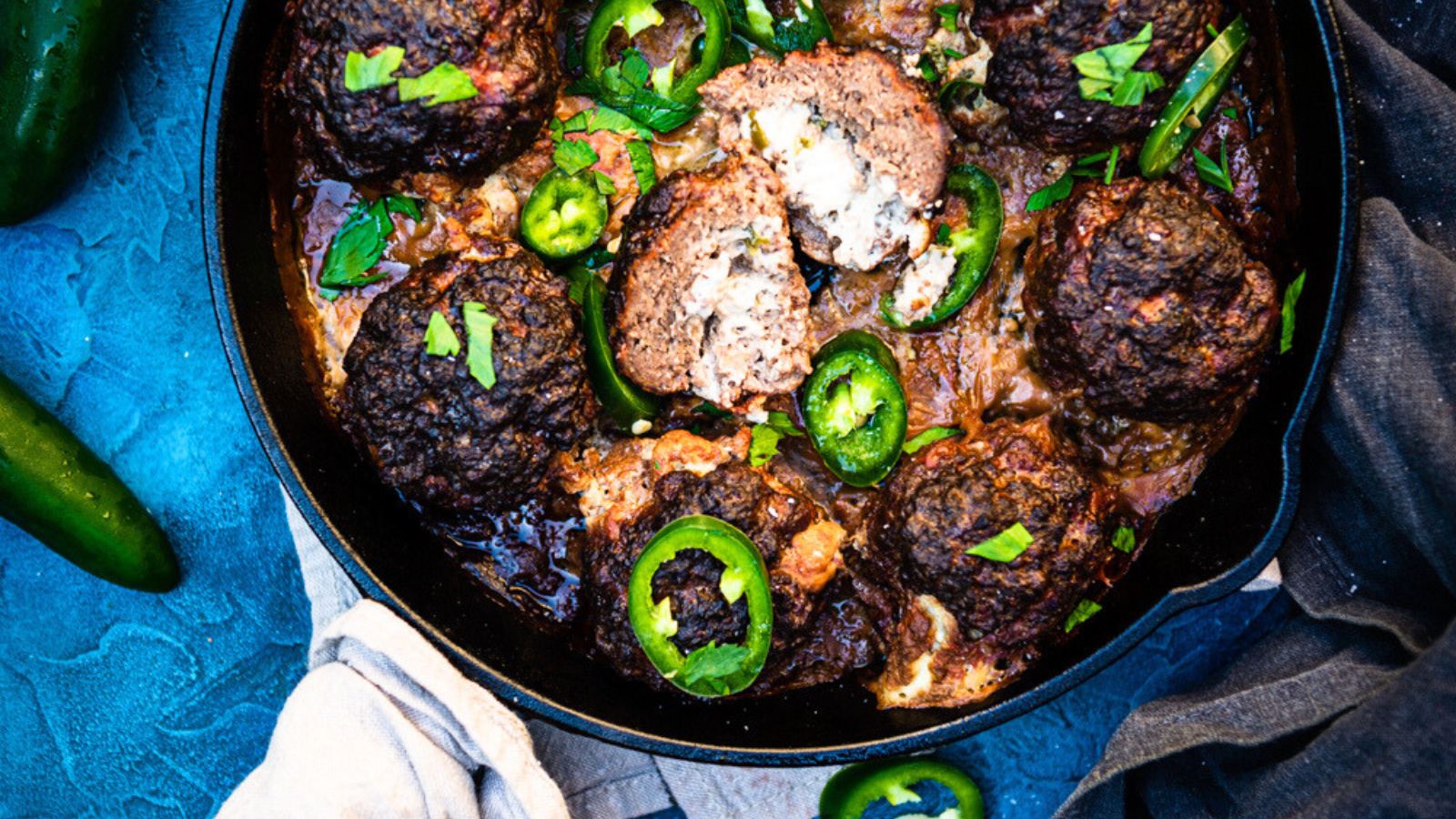 Meatballs in a pan with sliced jalapeños and garnished with herbs on a blue surface. Some meatballs are broken open.