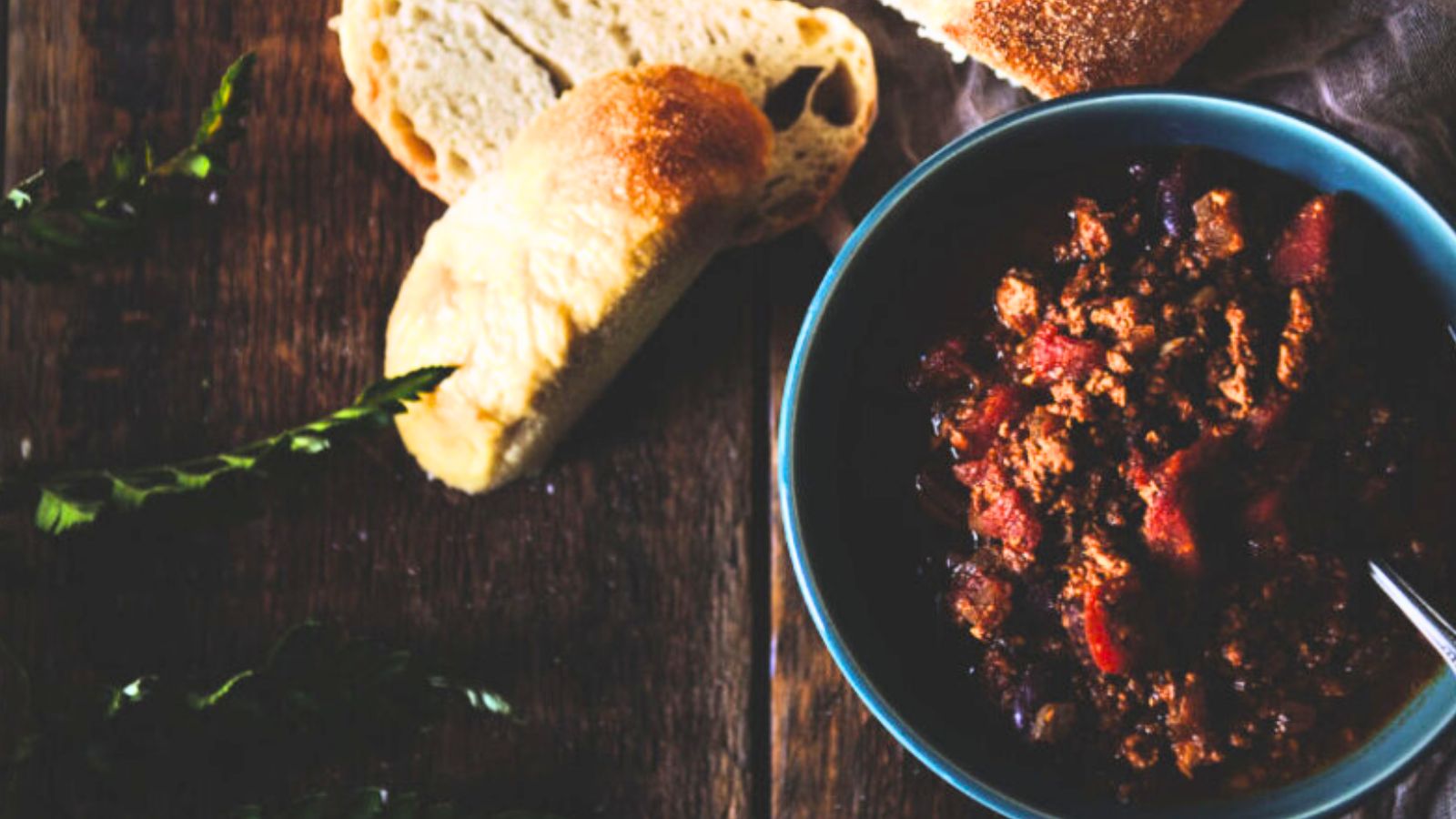 A bowl of chili, inspired by classic chili recipes, sits on a wooden table with a spoon, accompanied by sliced bread and fresh green leaves.