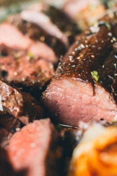 Close-up of a sliced sous vide venison tenderloin on a plate, garnished with herbs and accompanied by roasted garlic cloves.