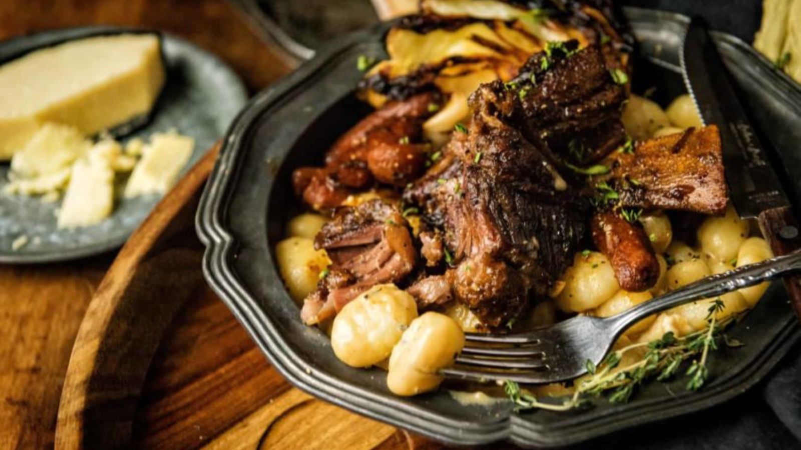 A plate of gnocchi topped with roasted beef and vegetables, garnished with herbs. A fork is placed on the plate, and a small plate of cheese is nearby.