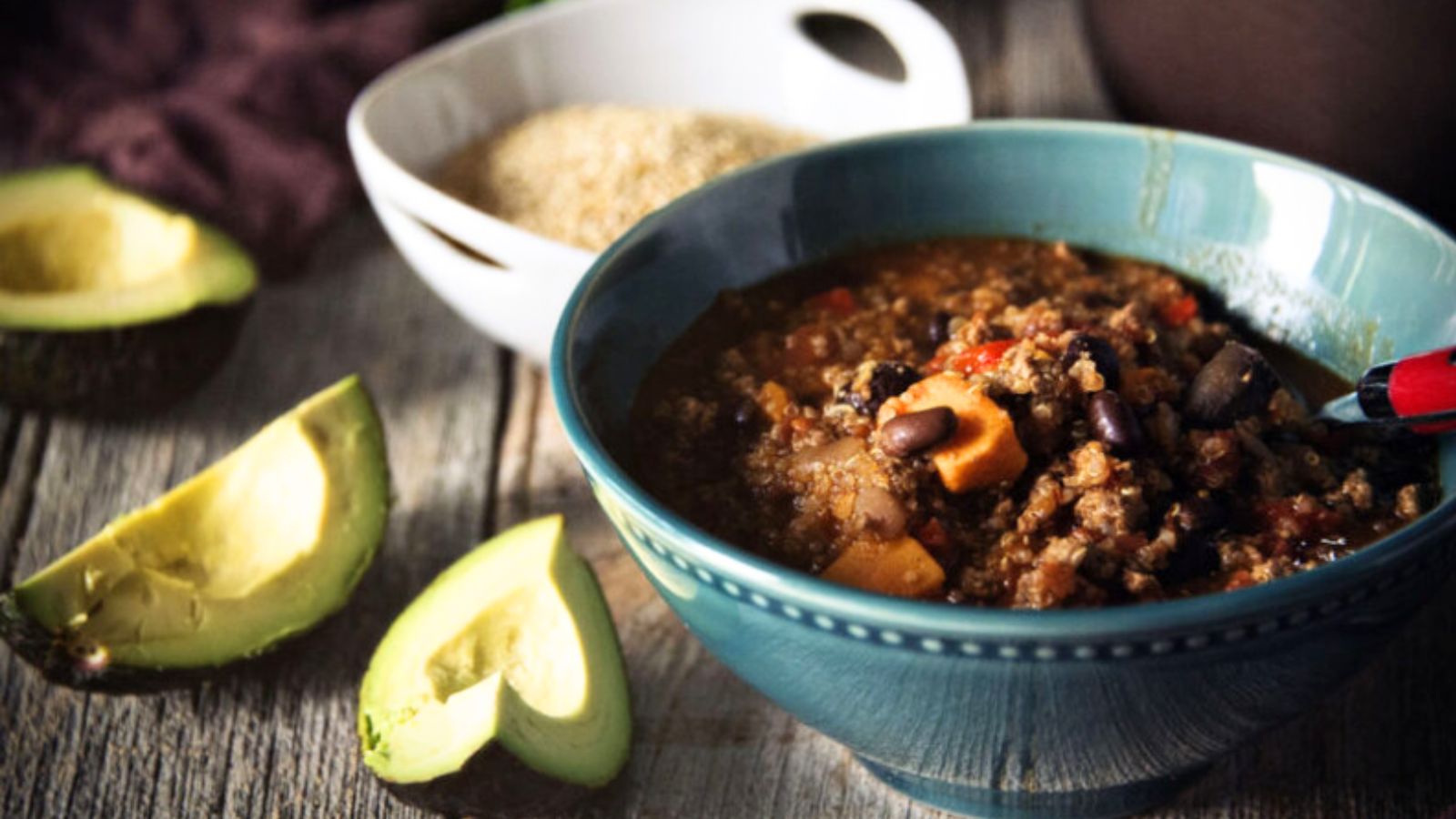 A delicious bowl of chili with beans, meat, and vegetables is on a table, embodying the essence of classic chili recipes. Next to it are creamy avocado slices and a bowl of wholesome grains, ready to be enjoyed.