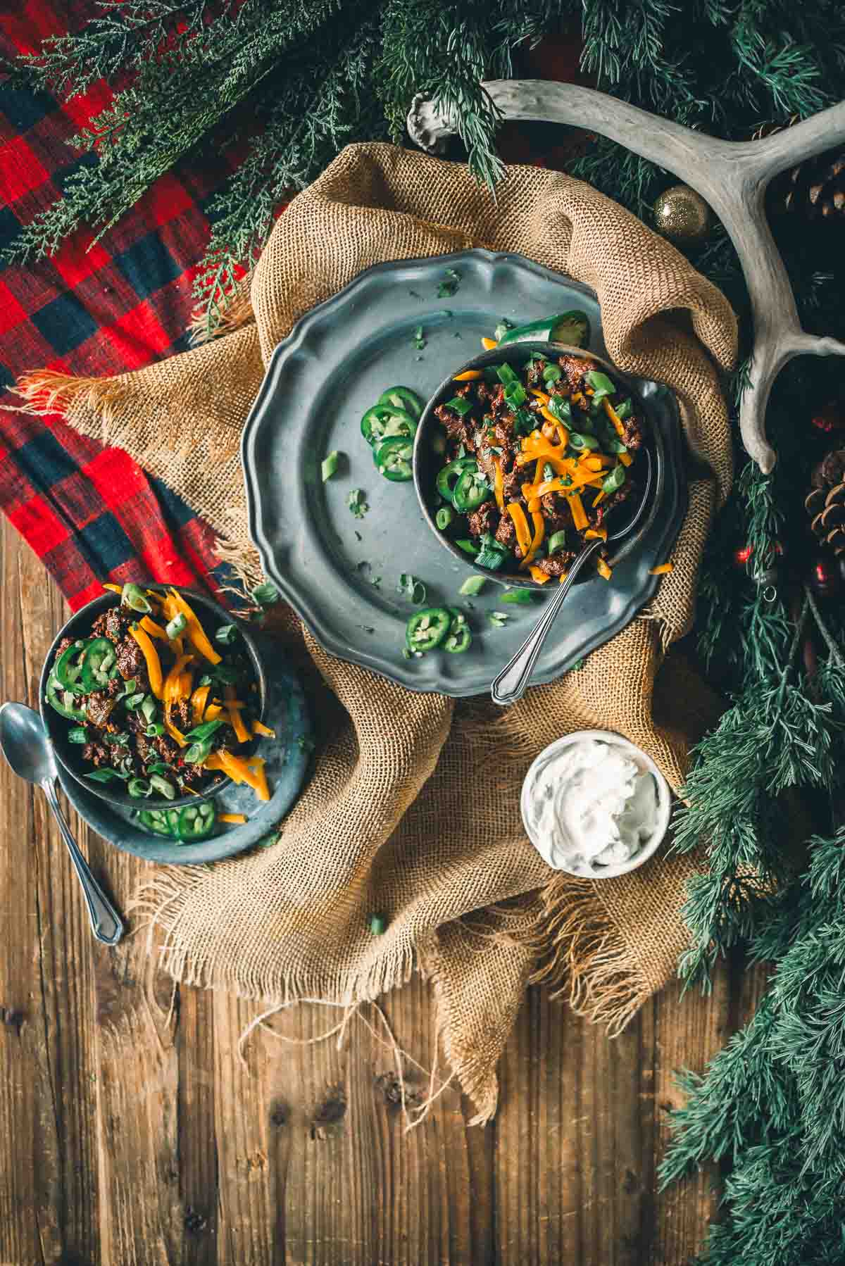 Two bowls of a deer meat chili topped with shredded cheese and herbs on a wooden table with burlap and plaid cloth underneath, surrounded by greenery and an antler.