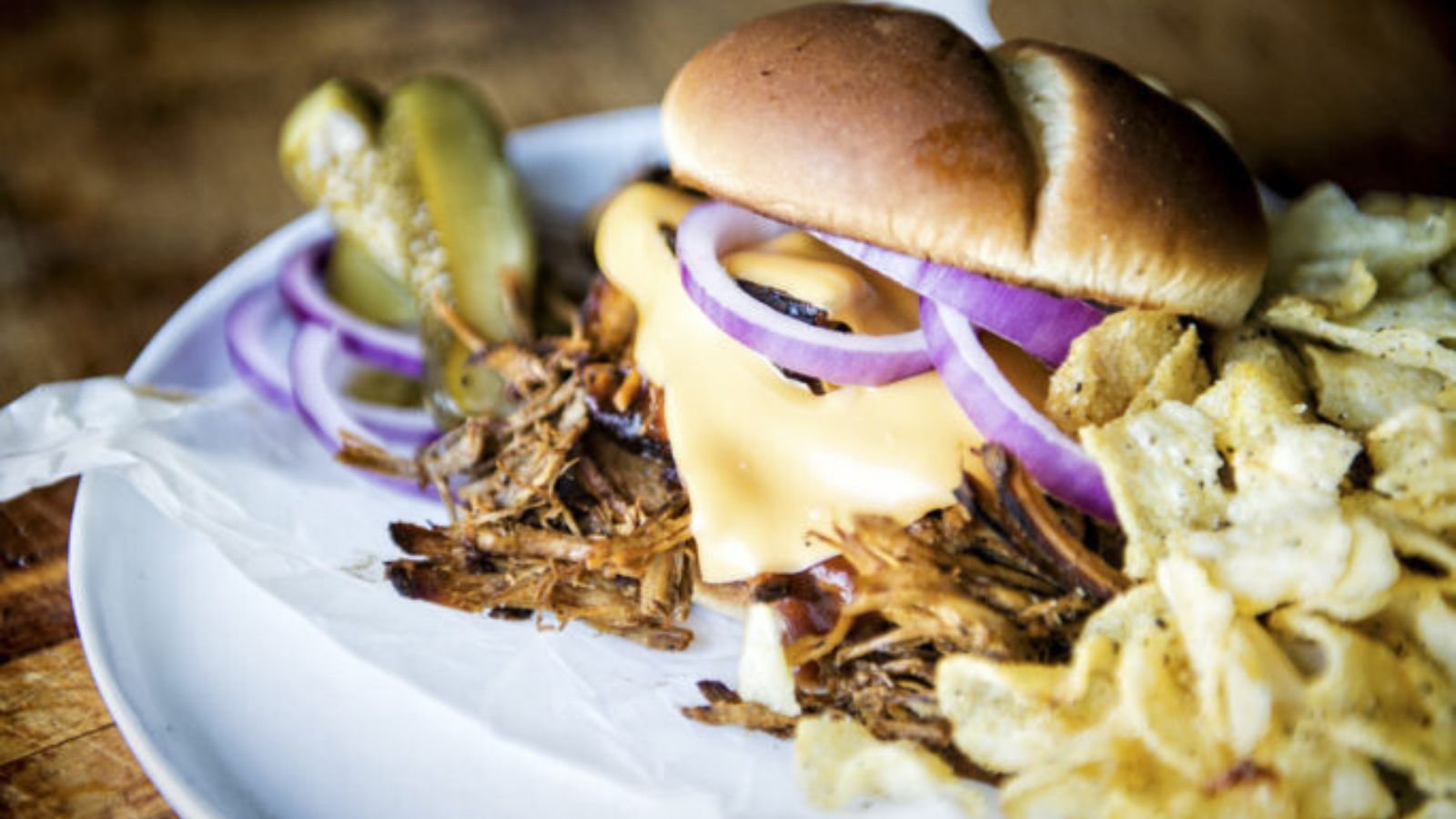 Pulled pork sandwich with melted cheese and red onions on a plate, served with pickles and potato chips.