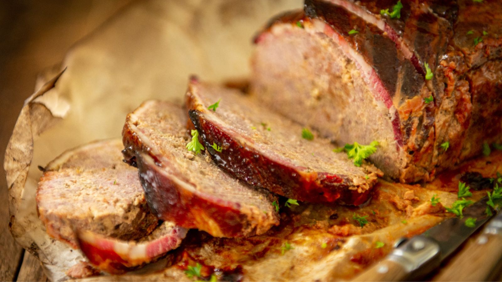 Sliced meatloaf with a dark crust is displayed on parchment paper, garnished with small green herbs. A knife is partially visible in the foreground.