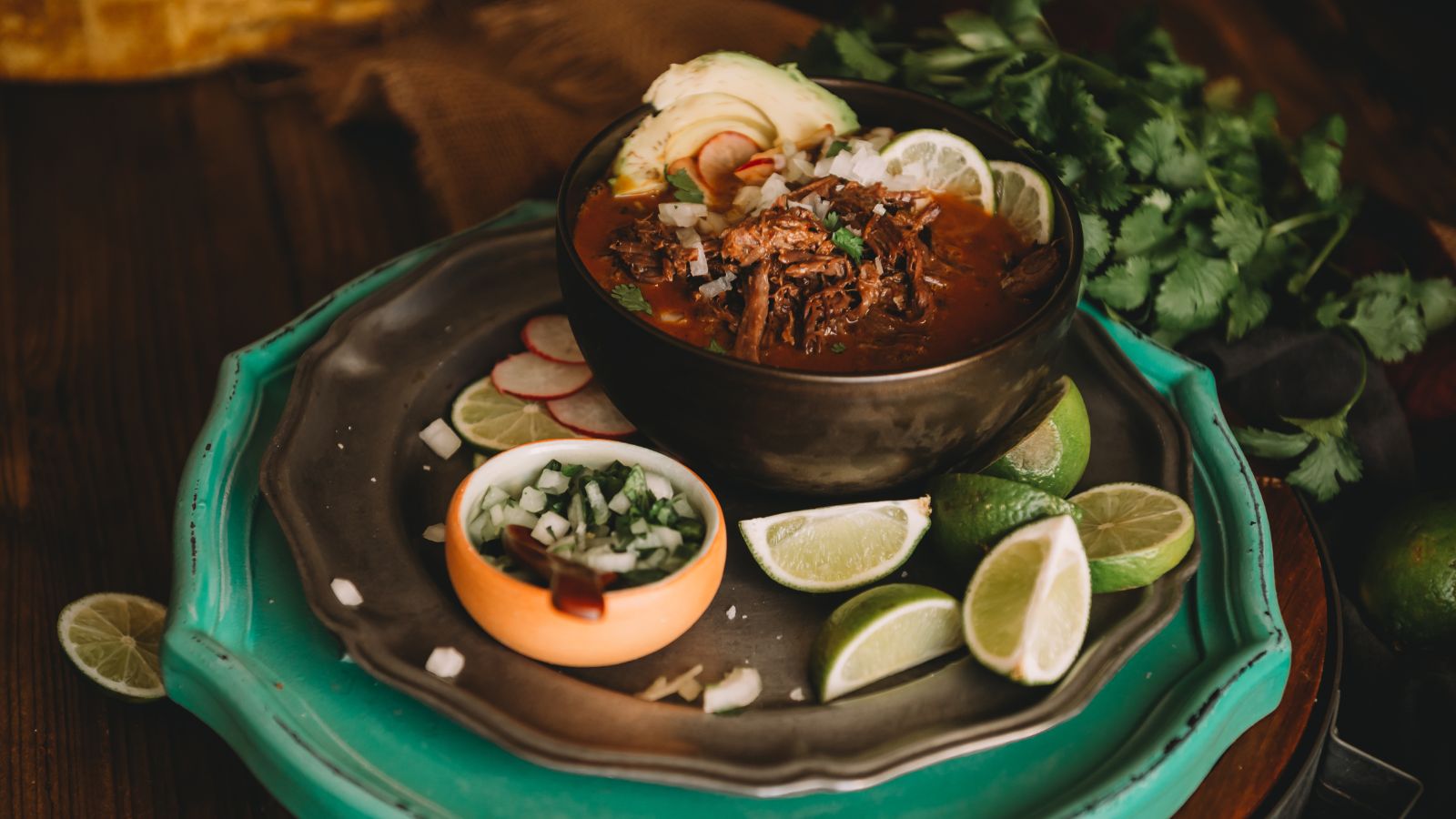 A bowl of beef stew garnished with avocado slices and radish, surrounded by lime wedges and cilantro on trays. A small dish with chopped onions and herbs is beside it.