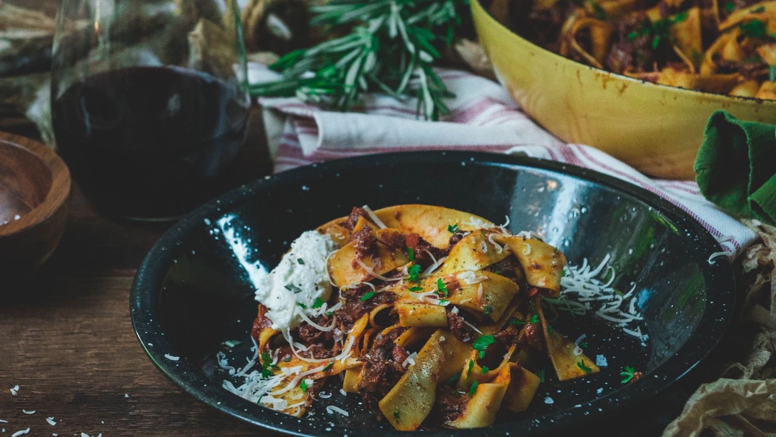 A plate of pappardelle pasta with meat sauce, garnished with grated cheese and herbs. A glass of red wine and a pan are in the background.