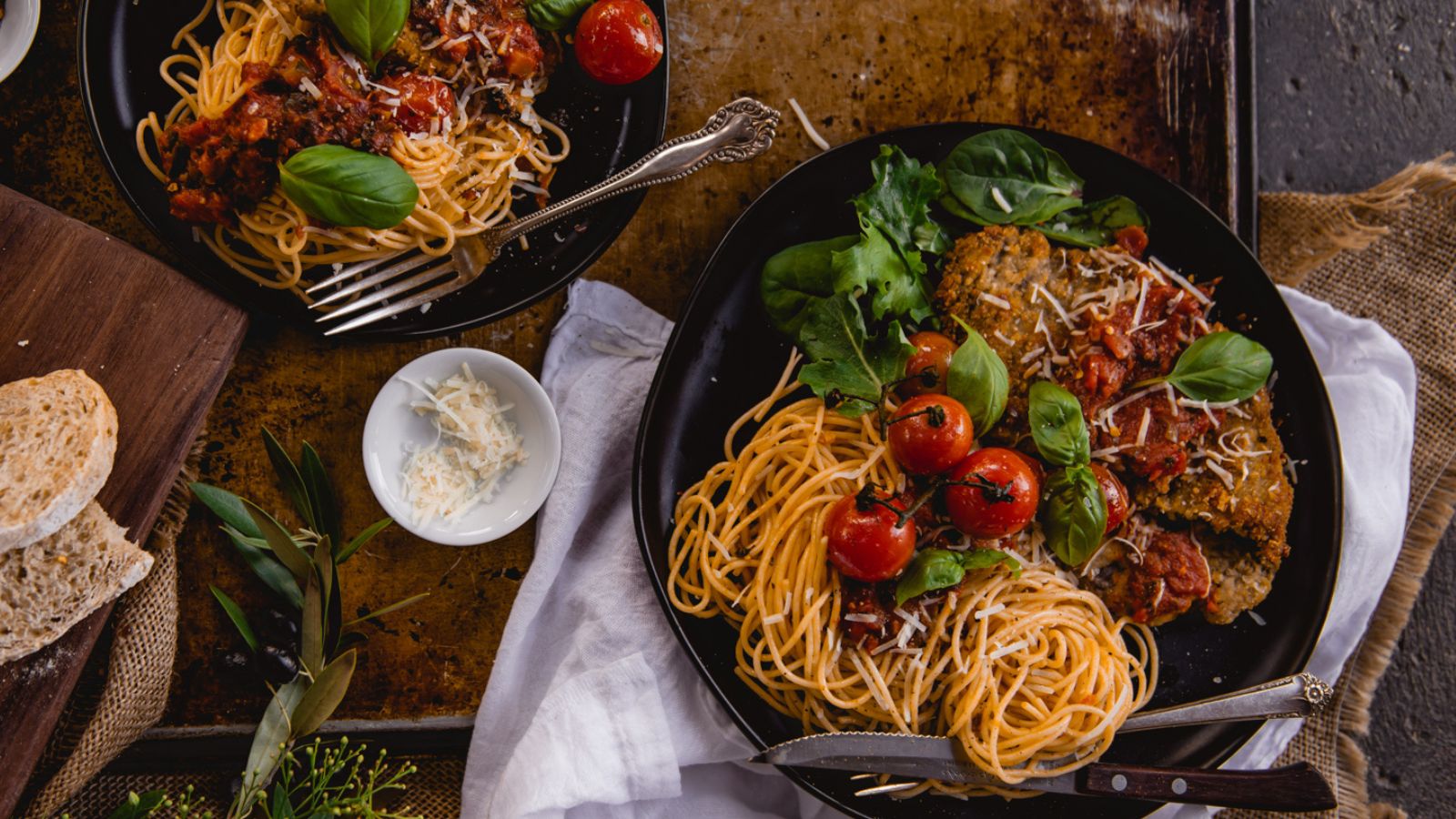 Two plates of spaghetti with meatballs, garnished with cherry tomatoes and basil. A small bowl of grated cheese and slices of bread are on the side.